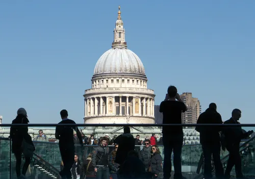 This urban architecture photograph was taken late in the morning during spring. The image prominently displays the Millennium Bridge in London, United Kingdom, with many people crossing the structure. St Paul's Cathedral, a well-known church and architectural landmark, is clearly visible in the background, its iconic dome dominating the skyline. The photograph captures the dynamic city atmosphere of London, showcasing both the bridge and the cathedral as significant elements of the city's architectural heritage. The scene emphasizes the connection between the modern Millennium Bridge and the historic St Paul's Cathedral, illustrating the blend of old and new found in this area of London.